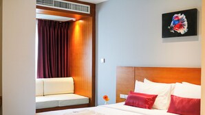 Una habitación de hotel con cama, una zona de estar con cortina, un cuadro en la pared y un pequeño jarrón con una flor.