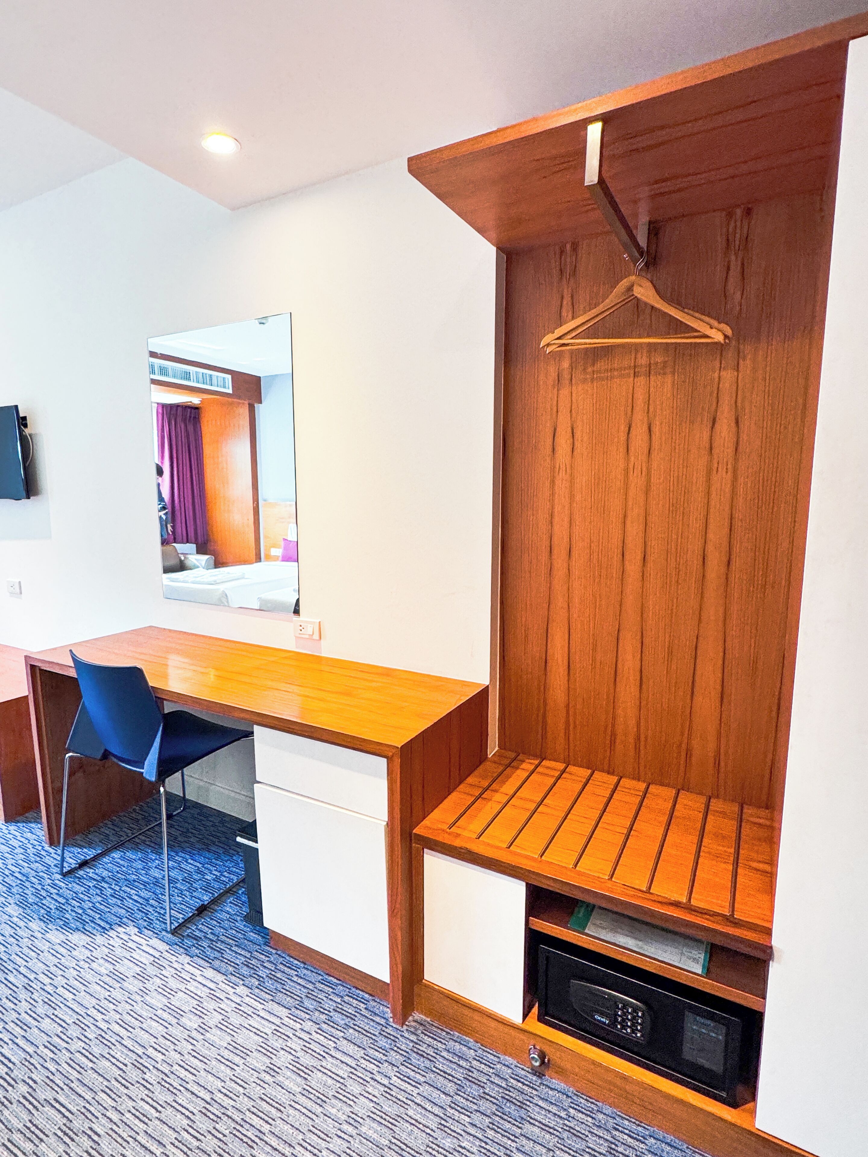 A hotel room with a wooden desk, a chair, a mirror, a hanging coat rack, and a built-in microwave.