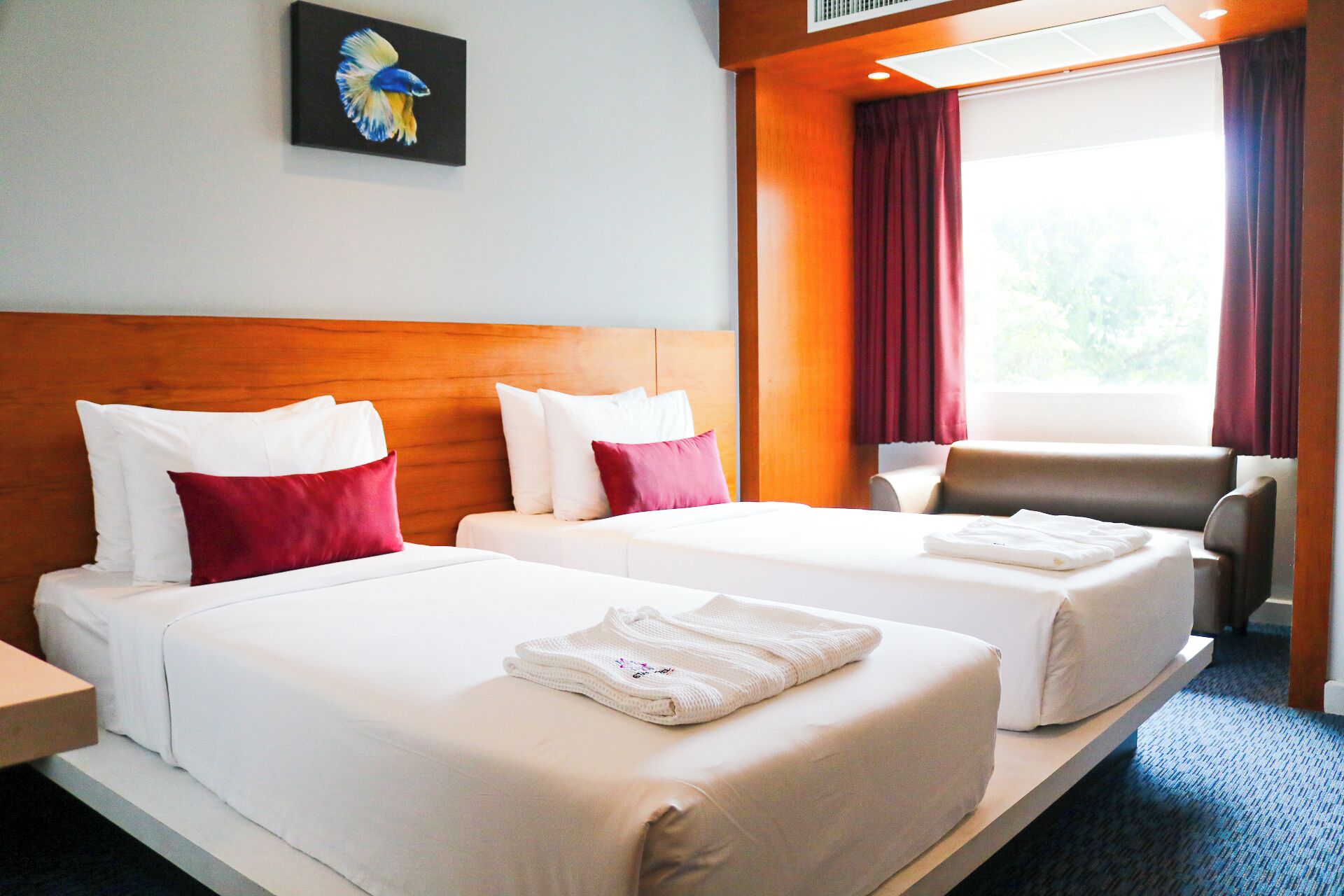 A hotel room with two beds, a wooden headboard, a painting, a window with curtains, and a chair.