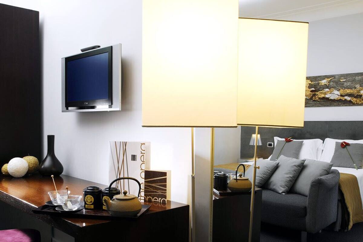 A modern hotel room with a flat-screen TV, a standing lamp, a sofa, and a bed with a headboard.
