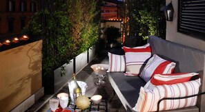 A cozy outdoor seating area with a sofa and cushions, a table with candles and a bottle, and a potted plant.