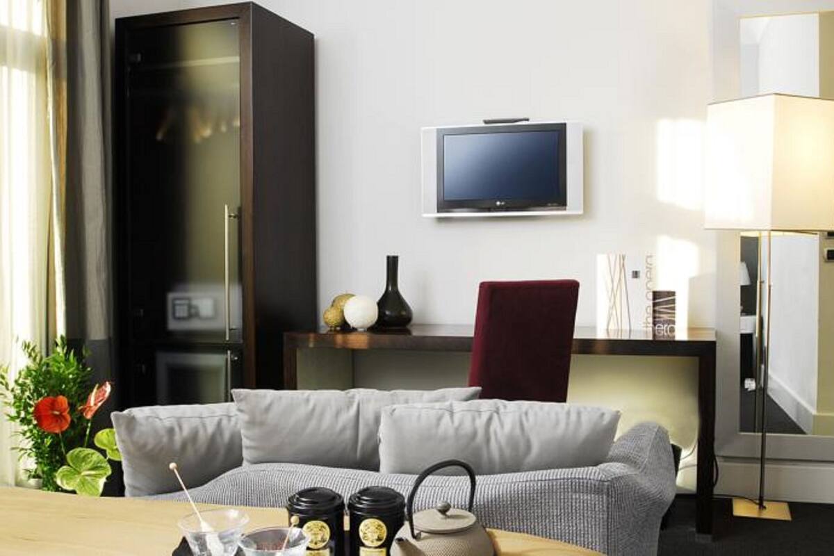 A modern hotel room with a grey sofa, a television on a stand, a desk with a lamp, and a large cabinet.