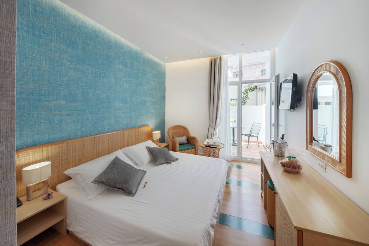 A hotel room with a large bed, a wooden nightstand, a wicker chair, a small table with a lamp, and a mirror on the wall.