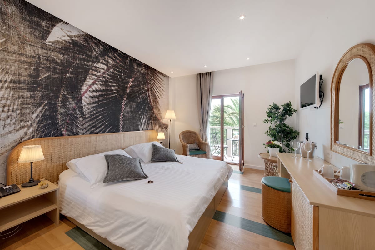A modern hotel room with a large bed, a desk with a laptop, a large mirror, and a balcony with a view of palm trees.