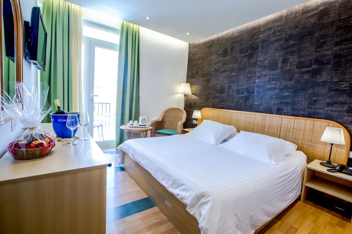 A hotel room with a large bed, a desk with a basket of fruit, a chair, a lamp, and a view of the outside.