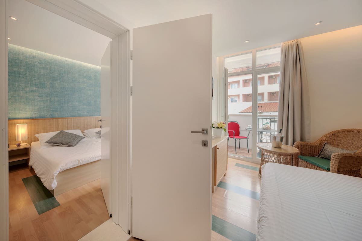 A hotel room with a bed, a desk, a wicker chair, and a balcony with a view of buildings.
