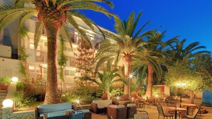 An outdoor patio area with tables and chairs, palm trees, and a building with balconies in the background. - Hotel Villa Adriatica - Adults only (Supetar)