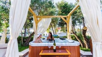 Un jacuzzi con un banco de madera y una botella de vino, rodeado de cortinas blancas y árboles.