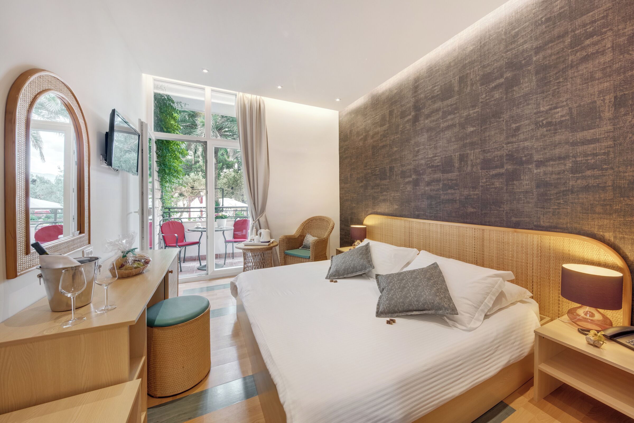 Una habitación de hotel moderna con una cama grande, un escritorio con lámpara, una silla de mimbre y vistas al exterior.