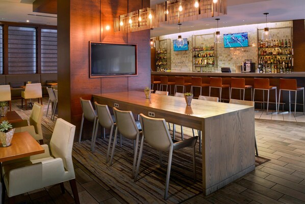 A modern bar area with a long counter, multiple bar stools, a flat-screen TV mounted on the wall, and a variety of bottles on shelves.