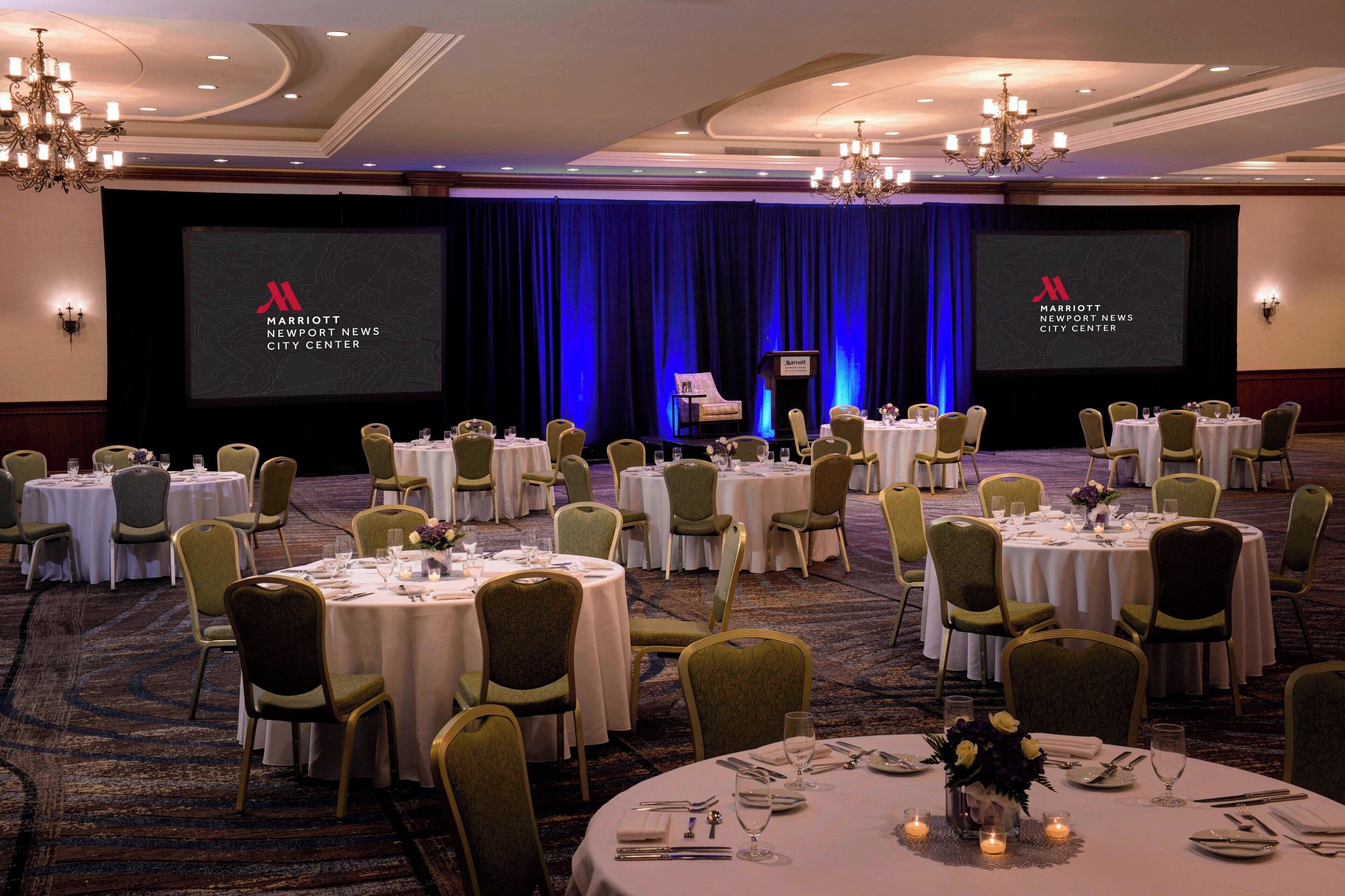 A conference room with multiple round tables set for a meeting, a stage with a screen displaying the Marriott logo, and a blue curtain backdrop.