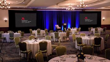 A conference room with multiple round tables set for a meeting, a stage with a screen displaying the Marriott logo, and a blue curtain backdrop.