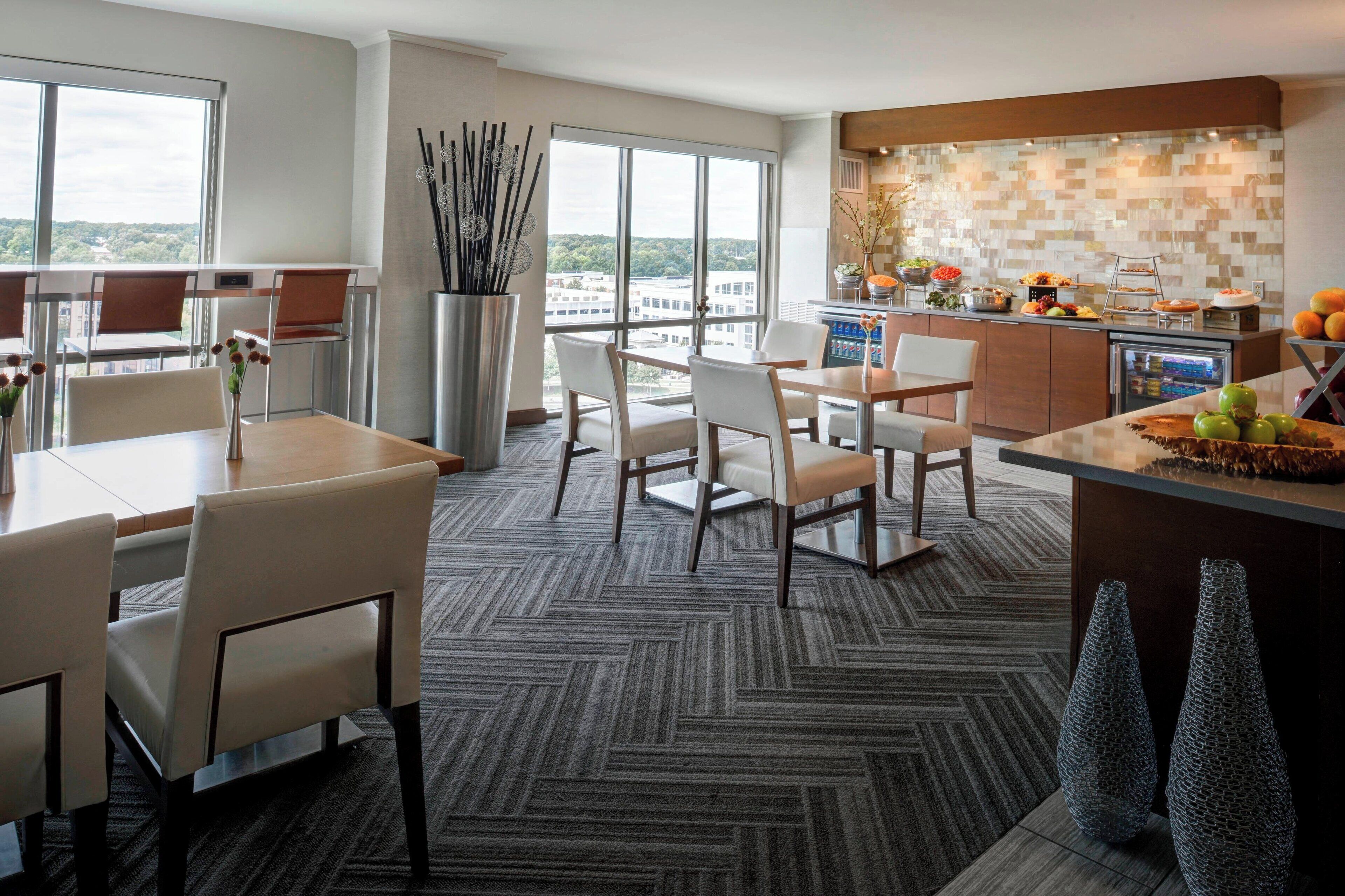 A modern hotel room with a dining area, a kitchen, and a large window offering a city view.
