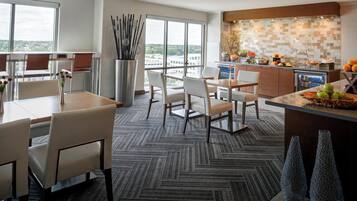 A modern hotel room with a dining area, a kitchen, and a large window offering a city view.