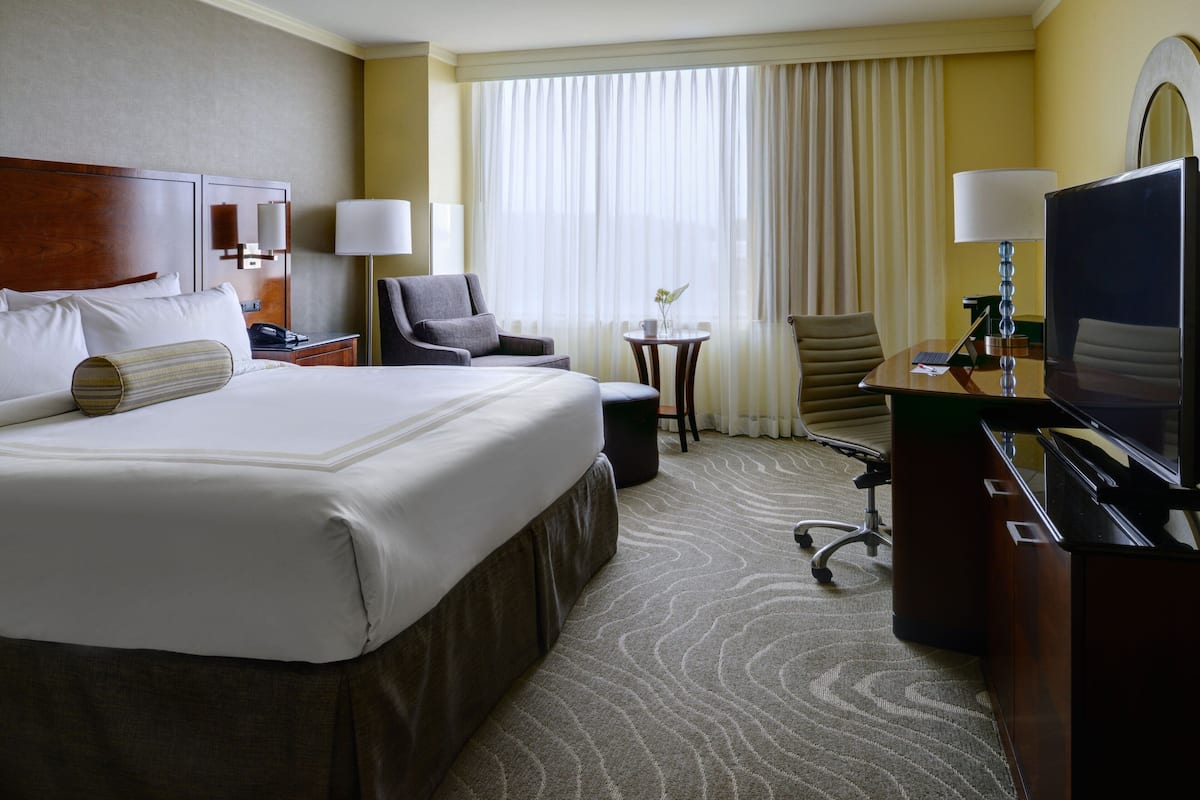 A hotel room with a large bed, a desk with a computer, a chair, a small table with a vase, and a television.