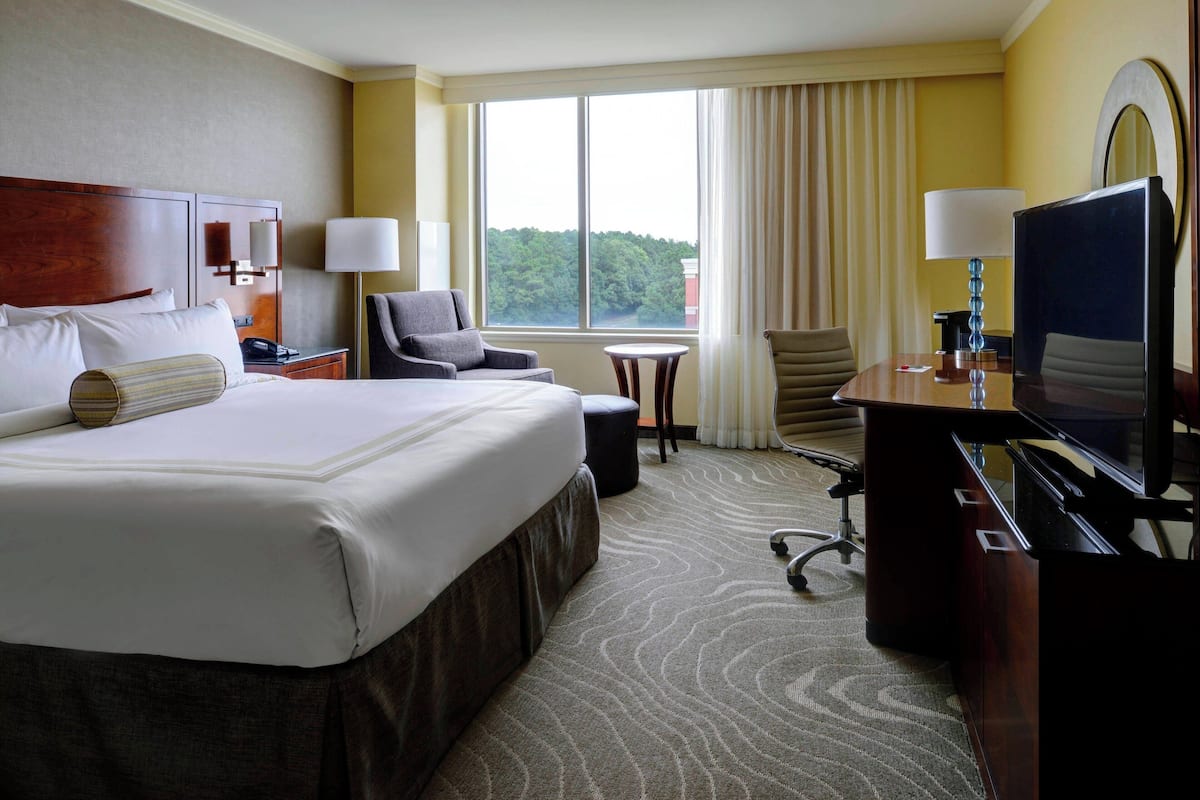 A hotel room with a large bed, two armchairs, a desk with a TV, and a window with curtains.