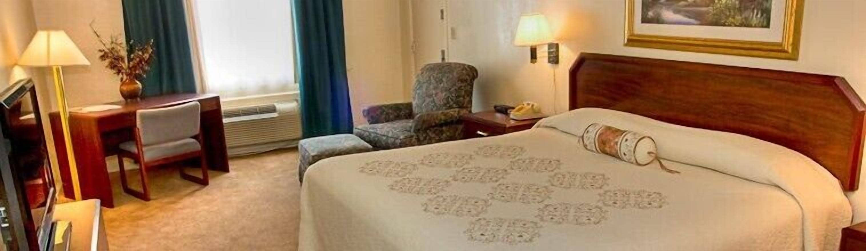A hotel room with a bed, a chair, a desk, a lamp, and a painting on the wall.