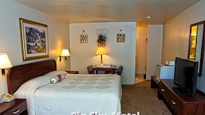 A hotel room with a bed, bedside table, lamp, desk, chair, television, and wall decor. - Big Sky Motel (Superior)