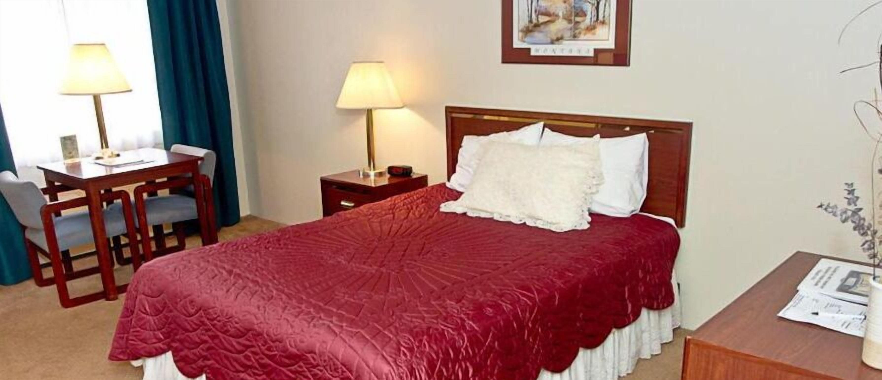 Habitación de hotel con cama, escritorio, silla, lámpara y un cuadro en la pared.