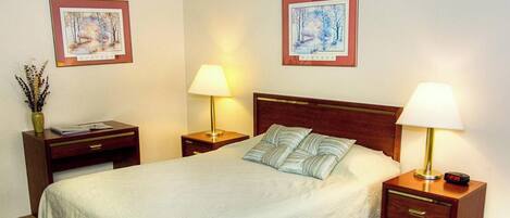 A hotel room with a bed, two bedside tables, lamps, a nightstand, a vase with dried flowers, and two framed pictures on the wall.