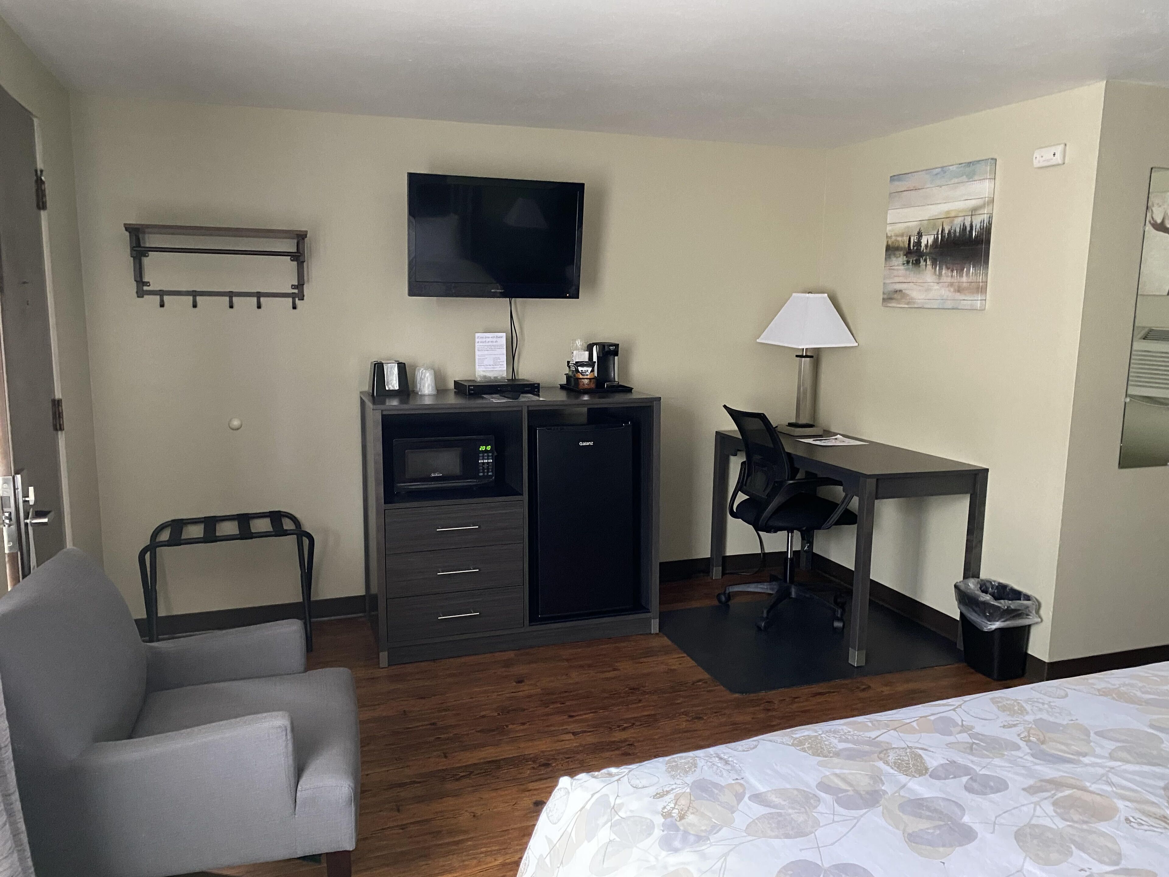 A hotel room with a bed, a desk with a chair, a TV, a microwave, a coffee maker, and a trash bin.