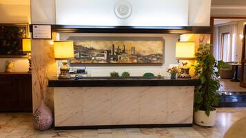 A reception desk with a cityscape painting above it, a vase with branches, and a potted plant to the right.