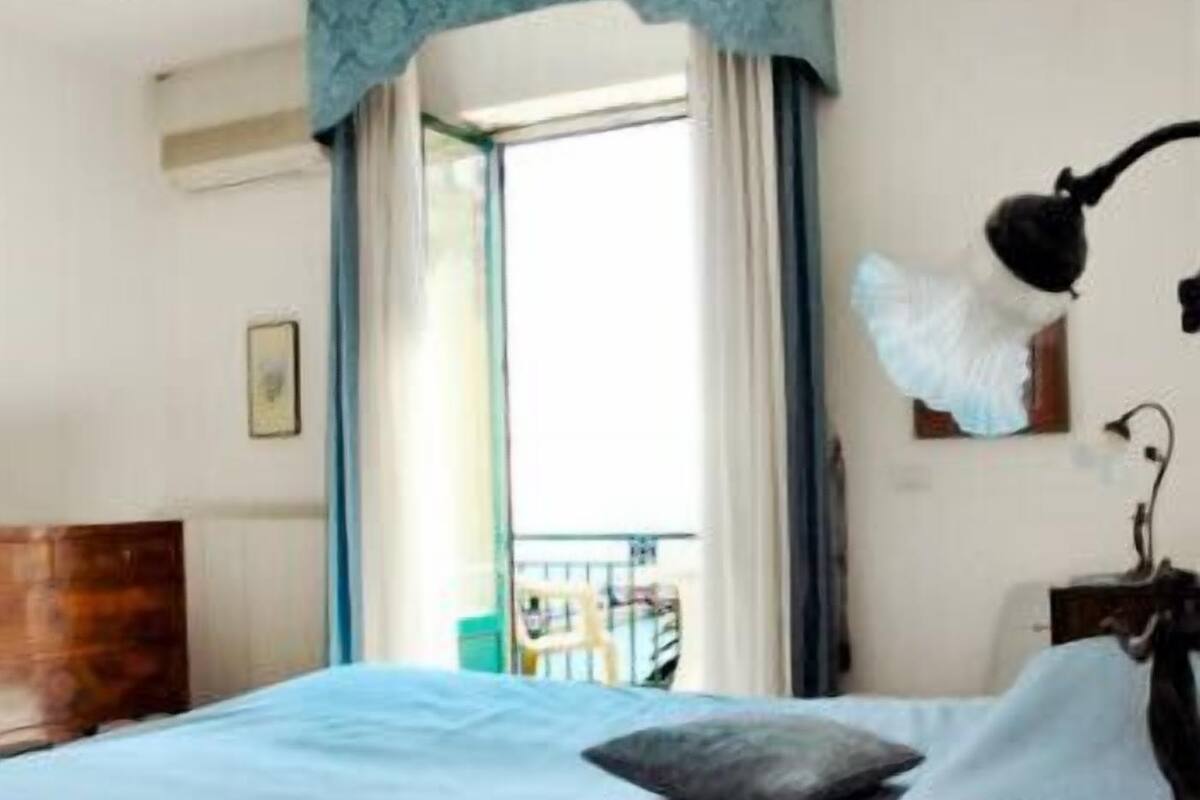 A hotel room with a bed, a nightstand, a lamp, and a view of the outside.