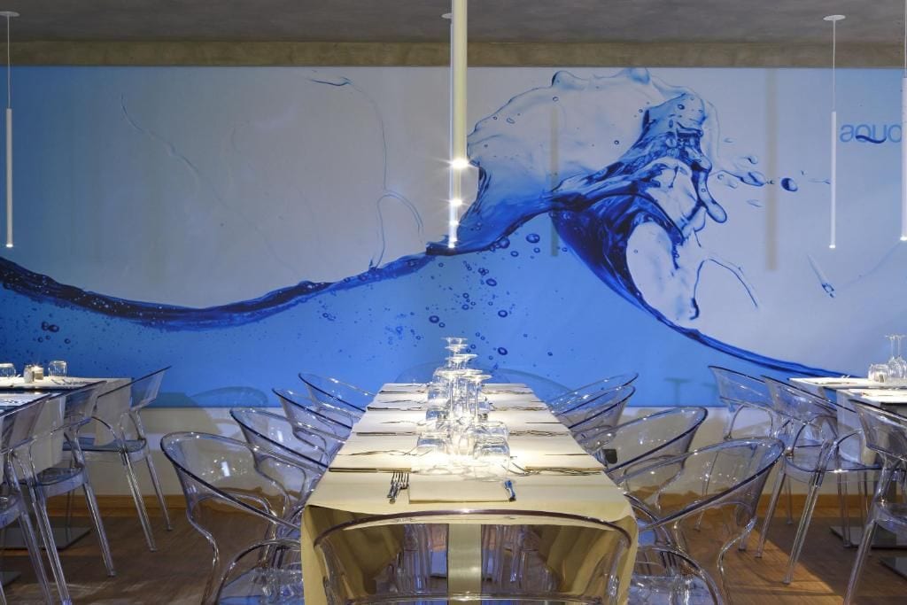A dining area with a long table set for a meal, clear chairs, and a large wall mural with a water splash design.