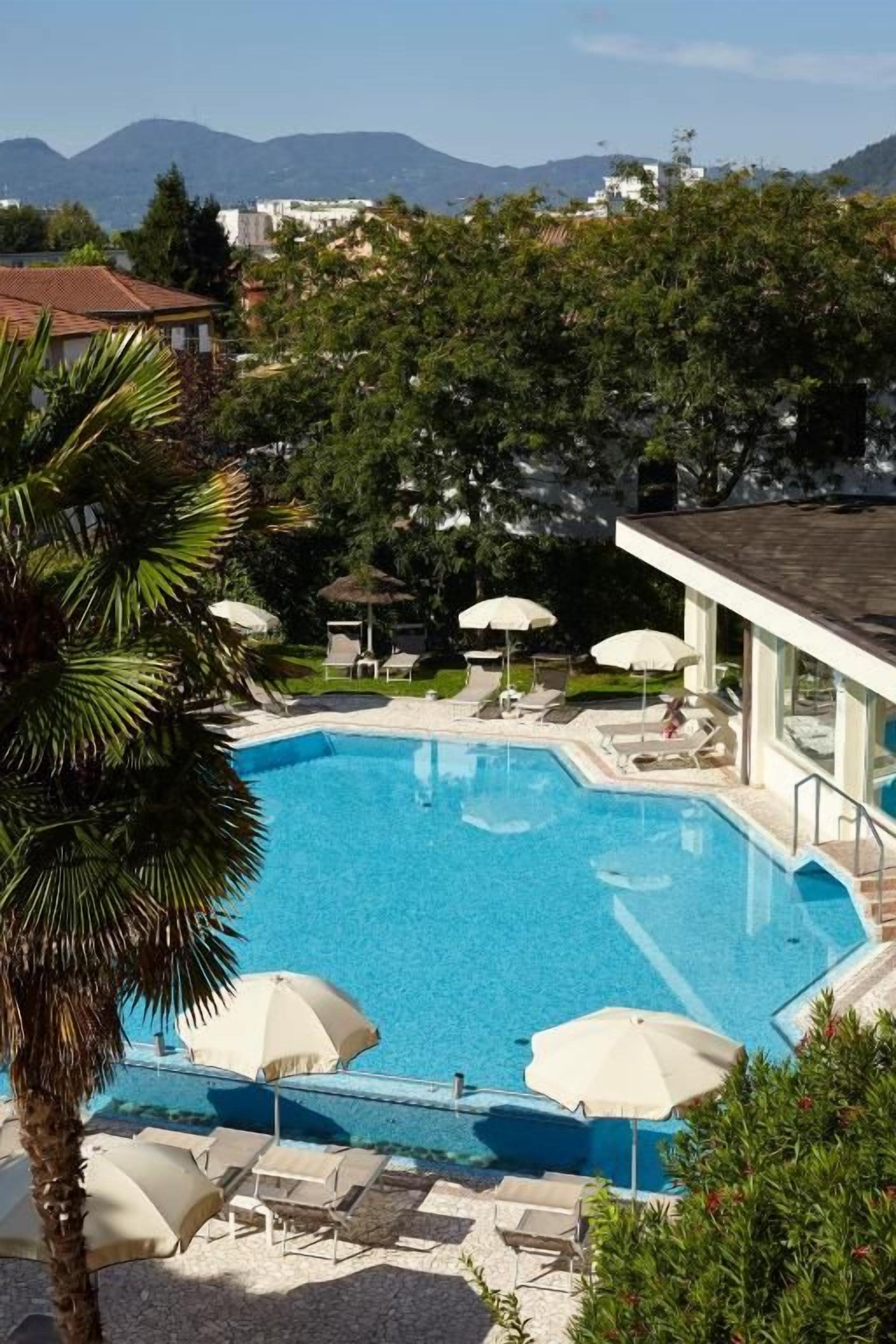 A swimming pool with lounge chairs and umbrellas, surrounded by greenery and buildings in the background.
