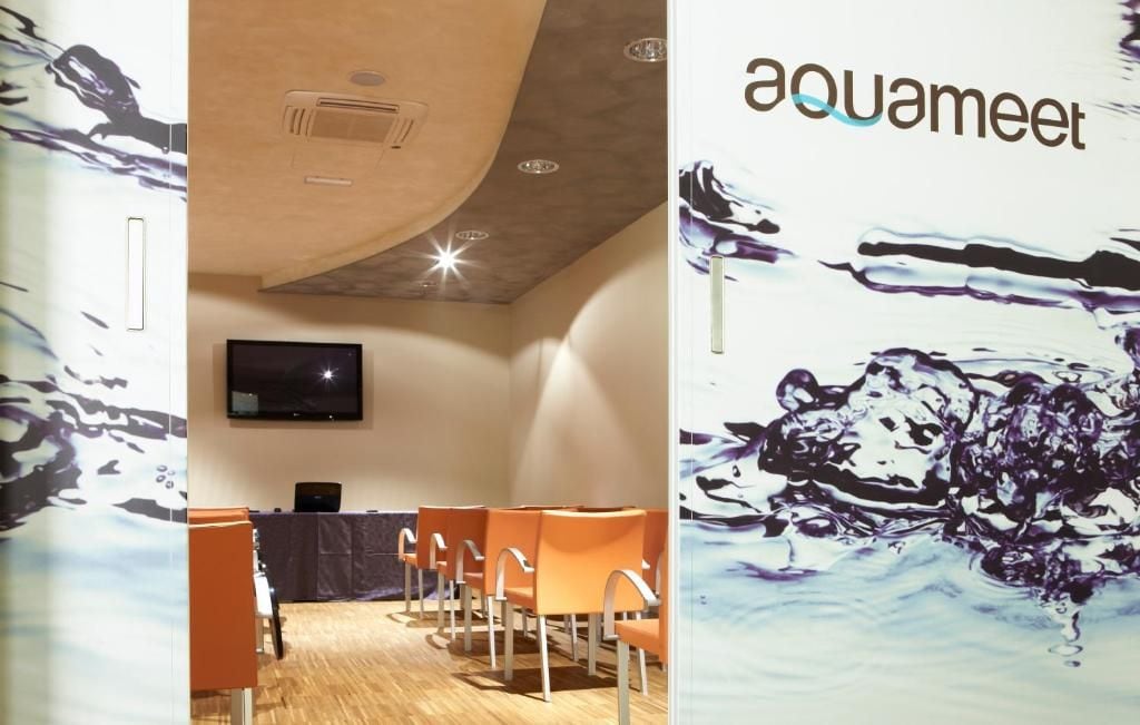Une pièce avec un téléviseur fixé au mur, une table avec des chaises et une grande affiche sur laquelle on peut lire le mot « aquameet ».