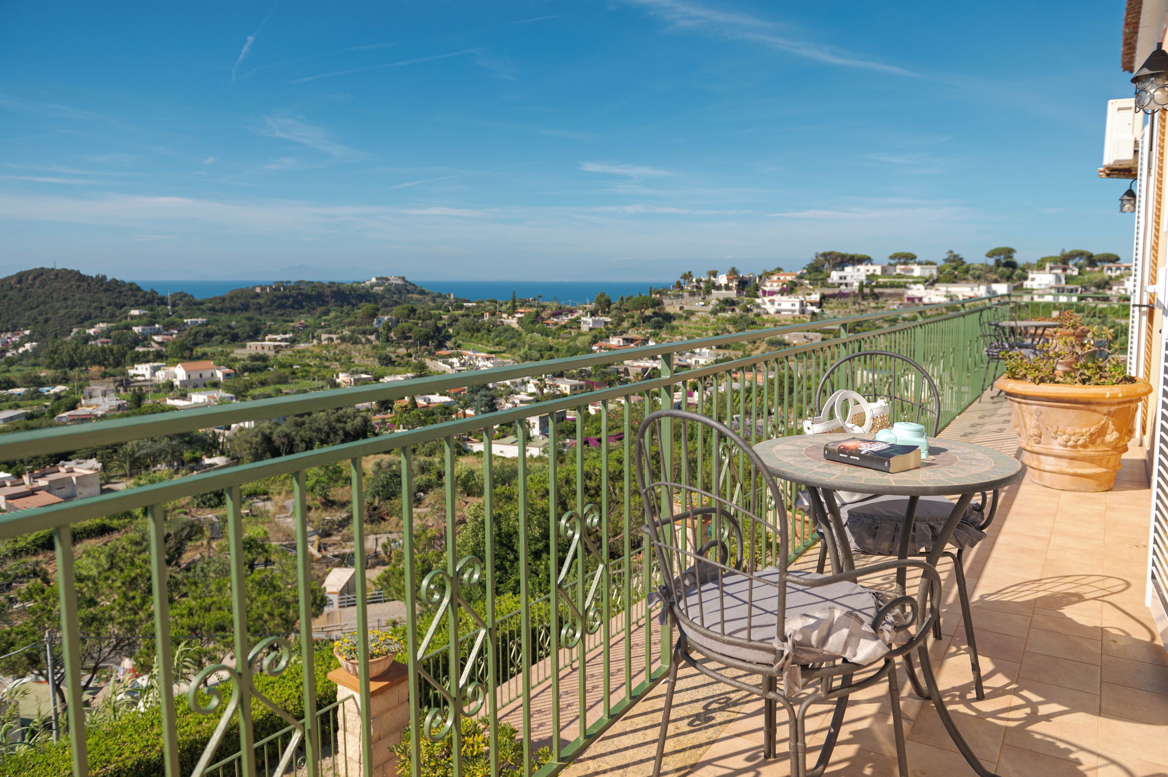 A balcony with a table and chairs, overlooking a coastal town with houses and greenery.