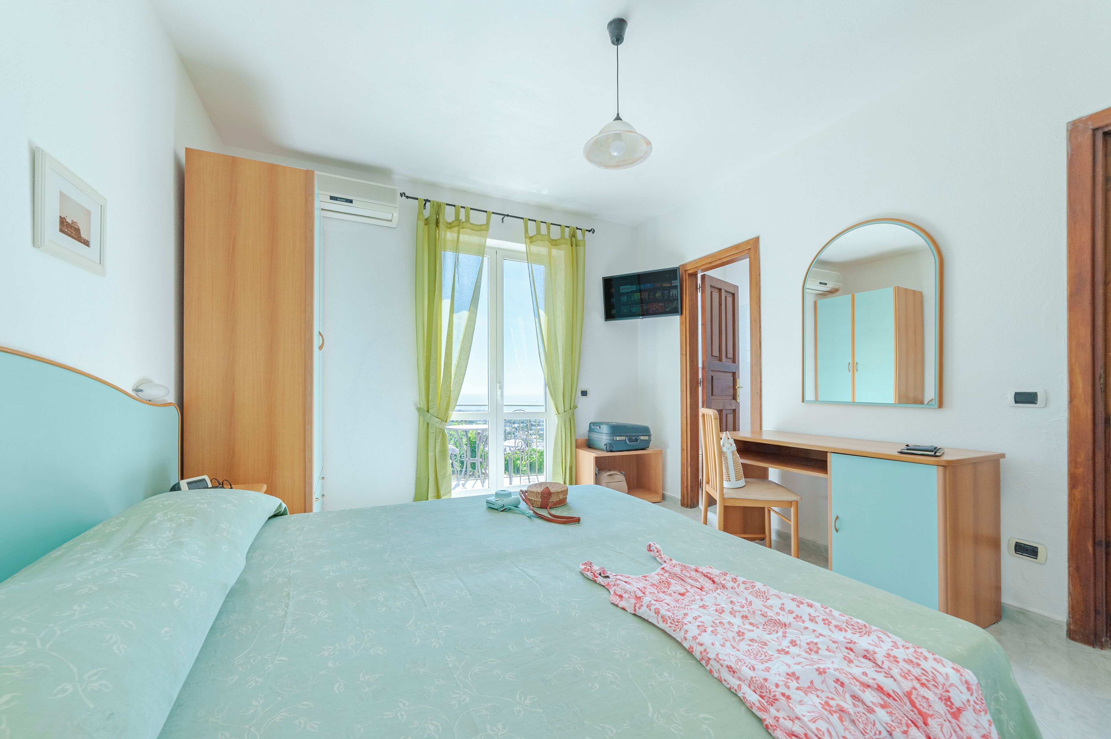 Un dormitorio con cama, escritorio, silla, armario, espejo y vistas al balcón.