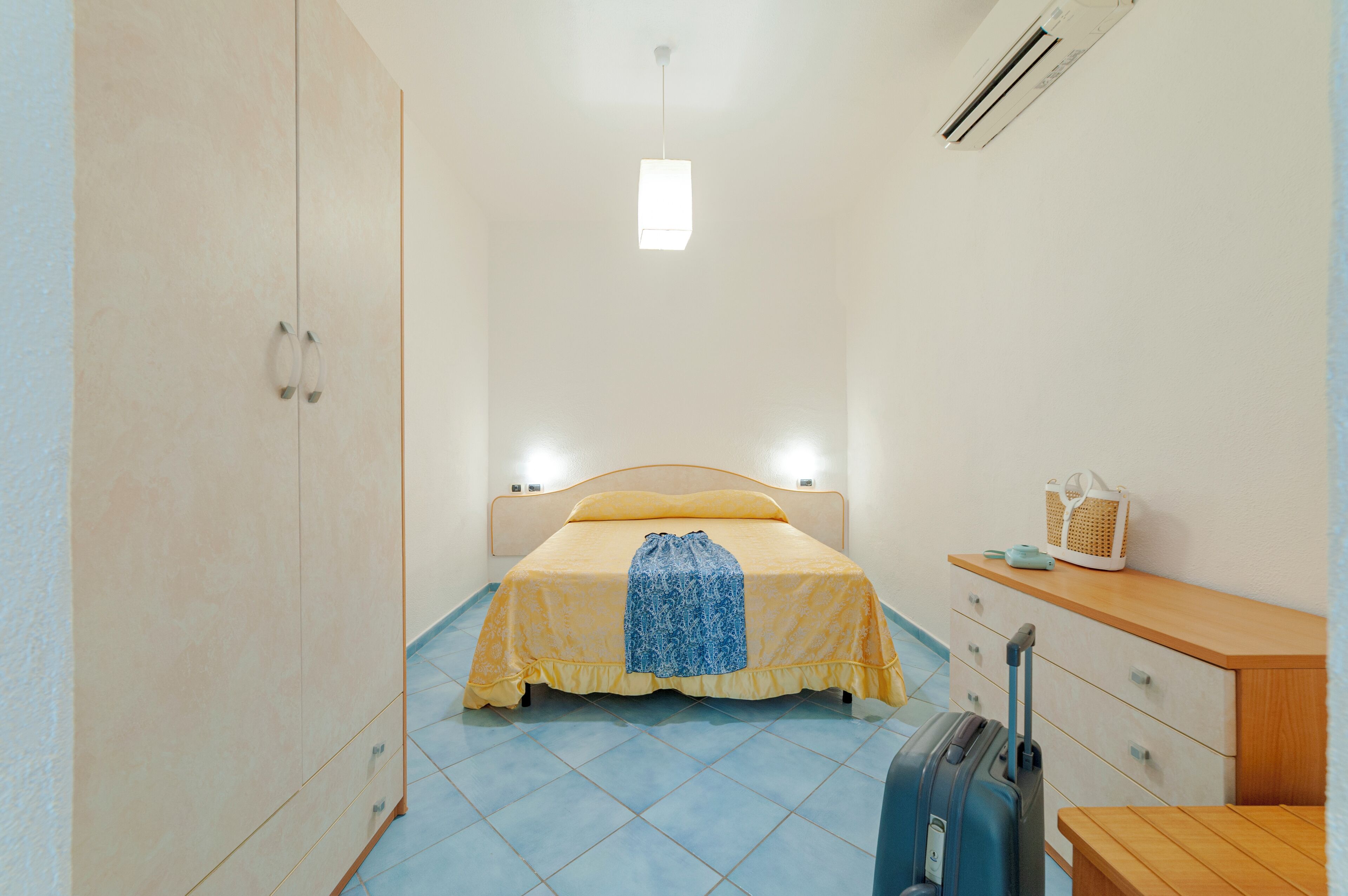 A small, bright room with a bed, a suitcase, a wooden dresser, and a wall-mounted heater.