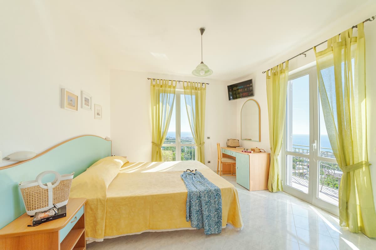 A hotel room with a bed, a desk, a chair, a TV, and a view of the sea.