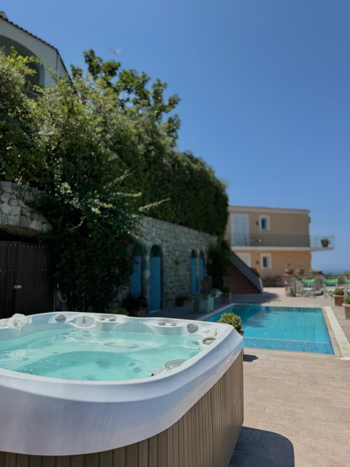 Un bain à remous avec une piscine en arrière-plan, entouré de verdure et bordé d’un mur de pierres.