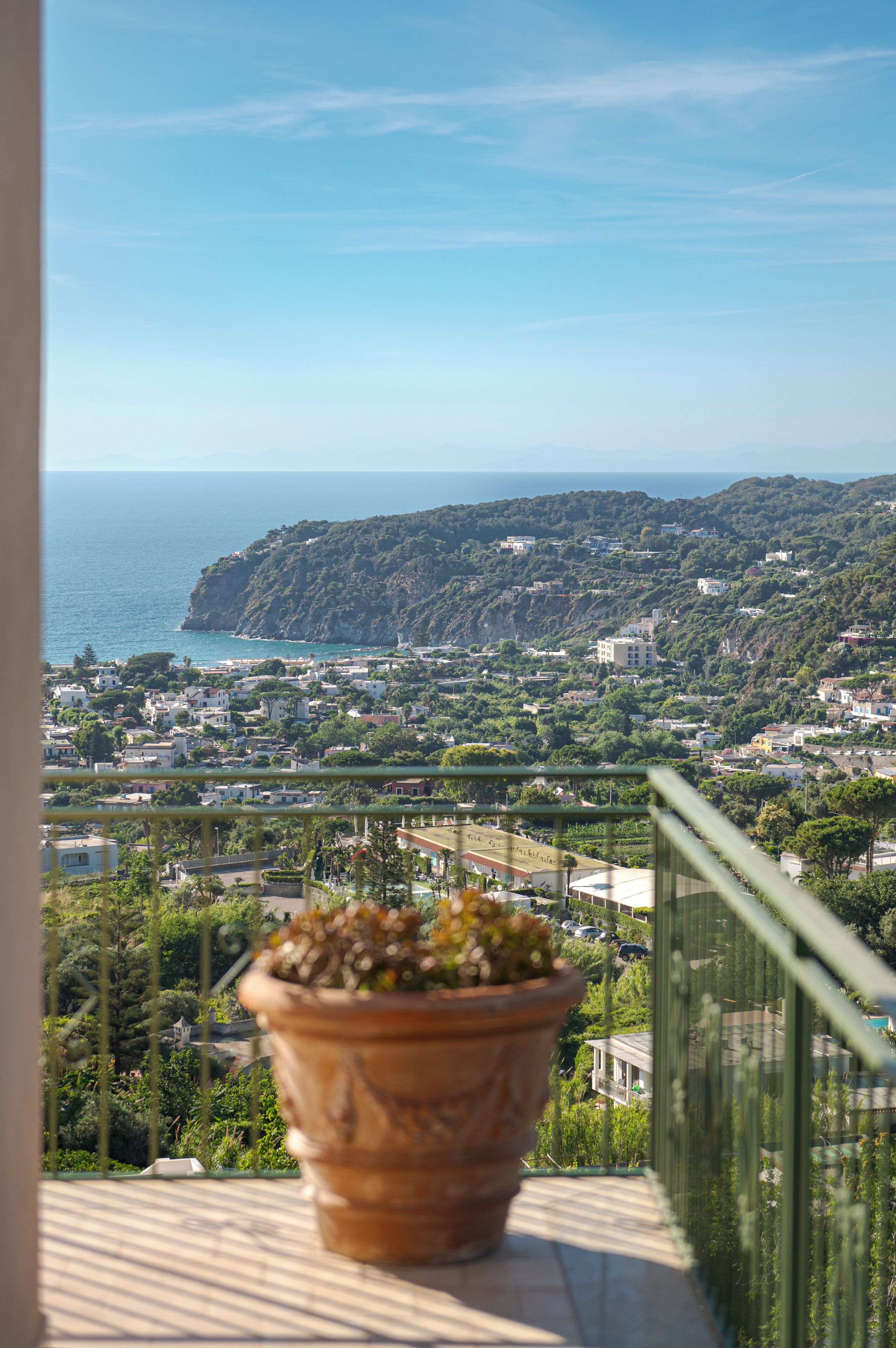 A balcony with a potted plant, overlooking a coastal town with buildings and greenery.