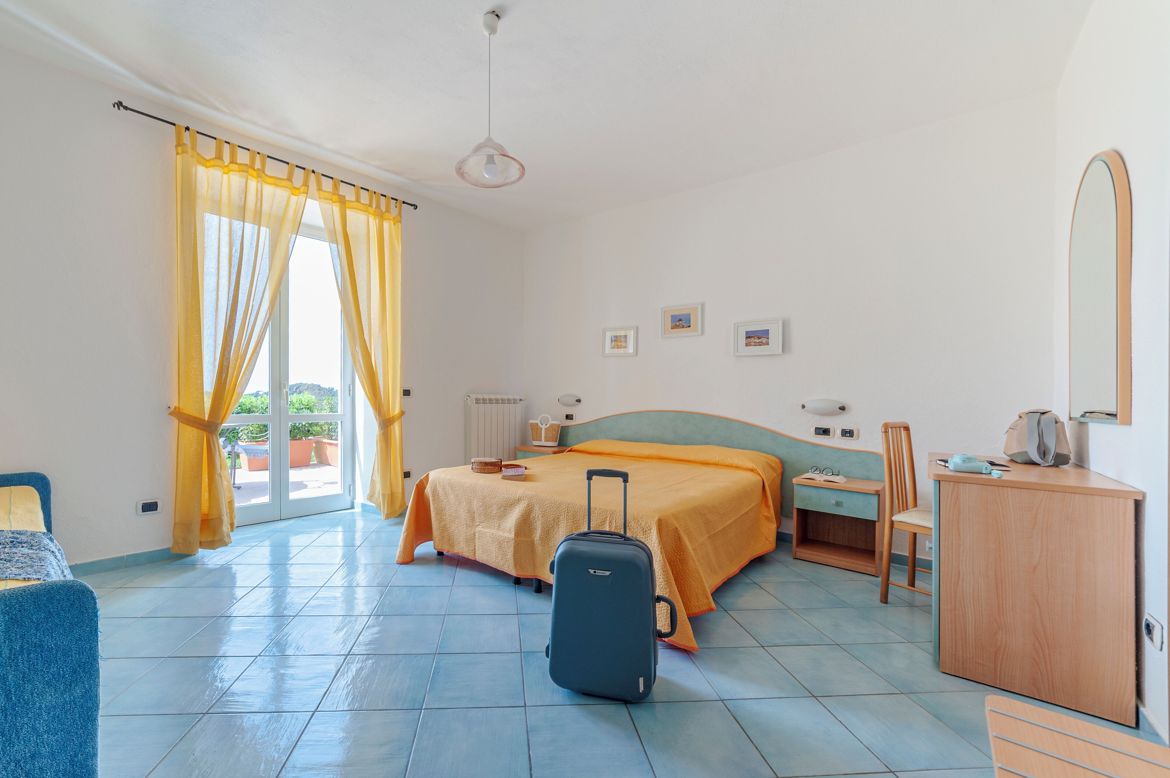 Une chambre d’hôtel avec un lit, un bureau, une chaise, une valise et une fenêtre avec des rideaux.