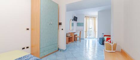 Une chambre comprenant un lit, une armoire, un bureau avec une chaise, une petite table avec un panier, et donnant sur un couloir où se trouvent des bagages.
