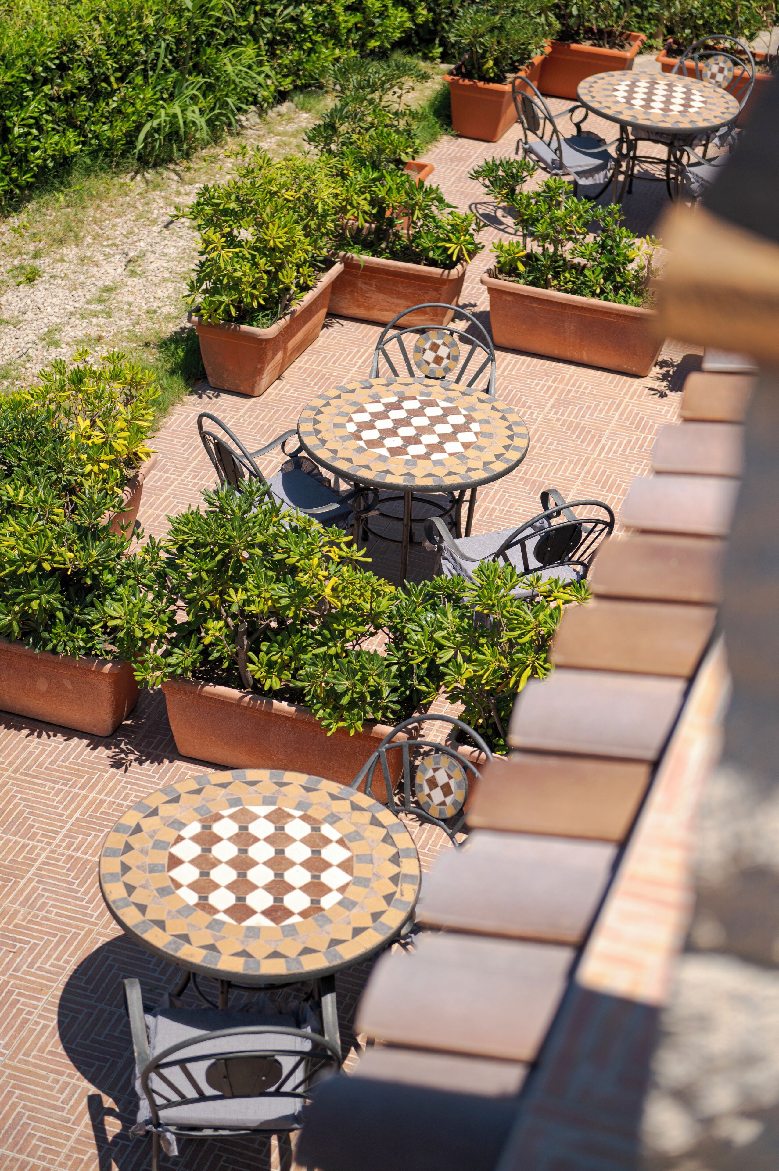 A rooftop terrace with multiple round tables and chairs, surrounded by potted plants and a patterned floor.