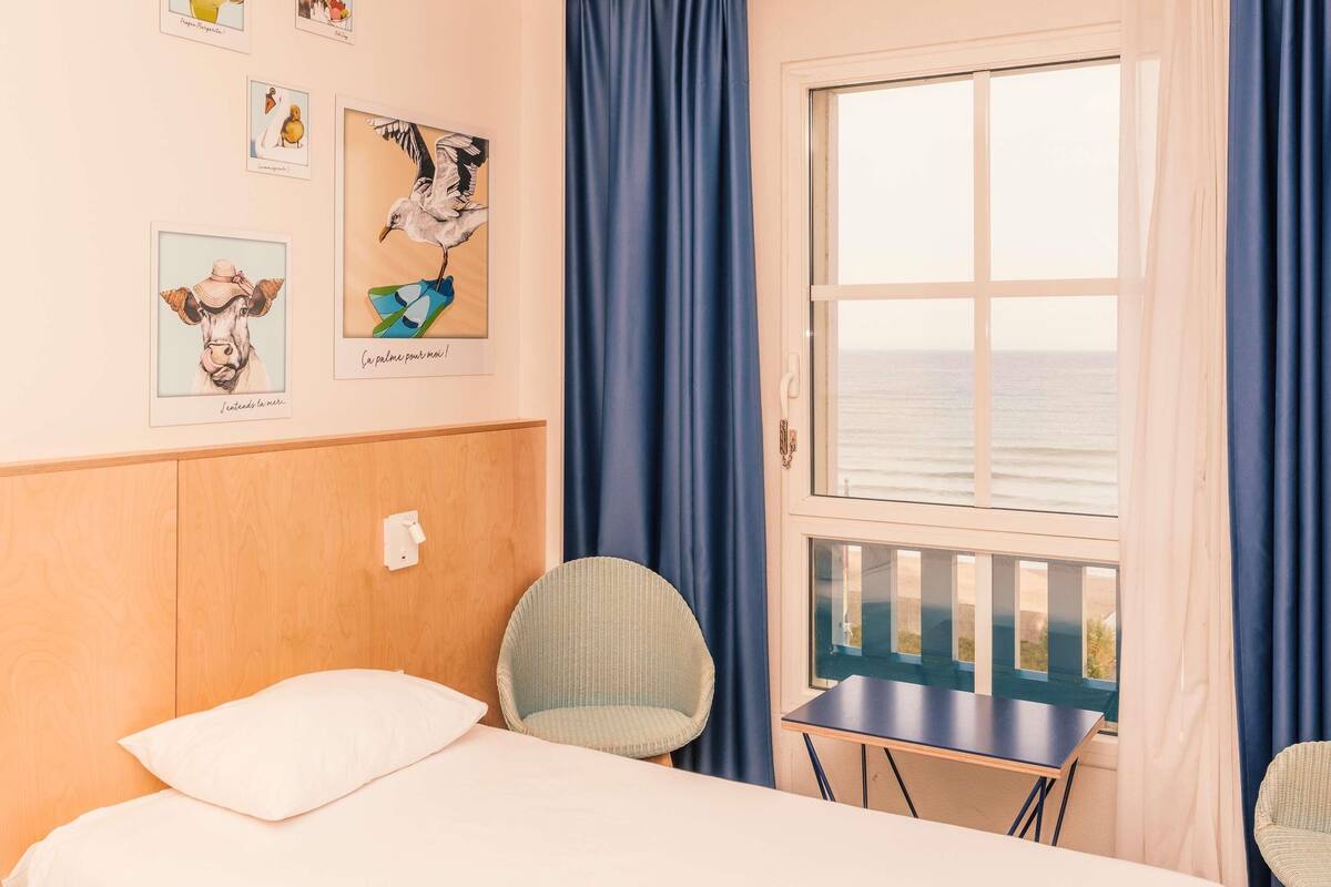 A room with a bed, a chair, a small table, and a window with a view of the sea.