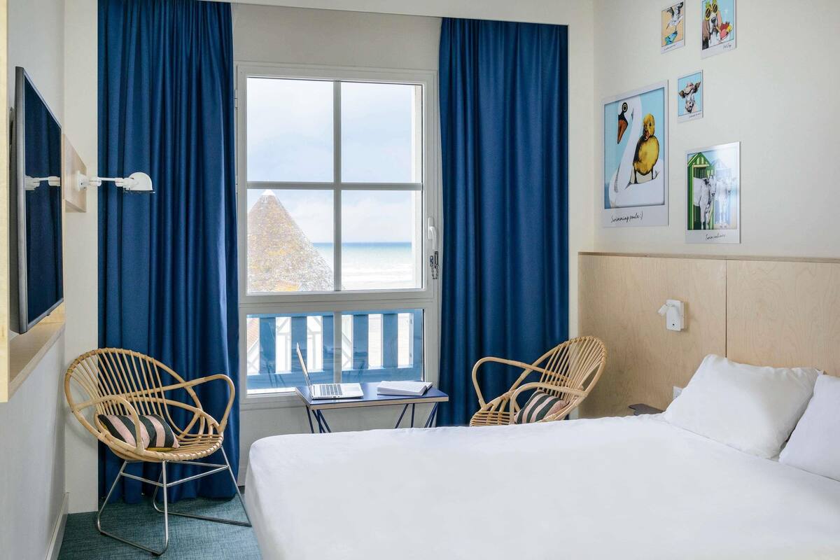 A hotel room with a bed, two wicker chairs, a small table, and a window with a view of the sea.