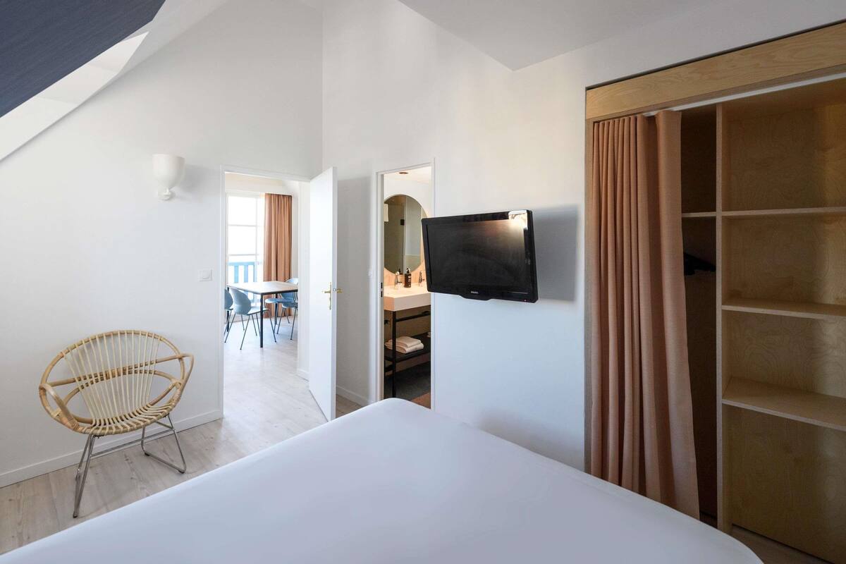 A hotel room with a bed, a television mounted on the wall, a wooden wardrobe, and a wicker chair.