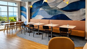 A modern restaurant interior with a wave-patterned mural, wooden tables, and a view of the outdoors. - Hotel ibis Styles Deauville Villers Plage (Villers-sur-Mer)