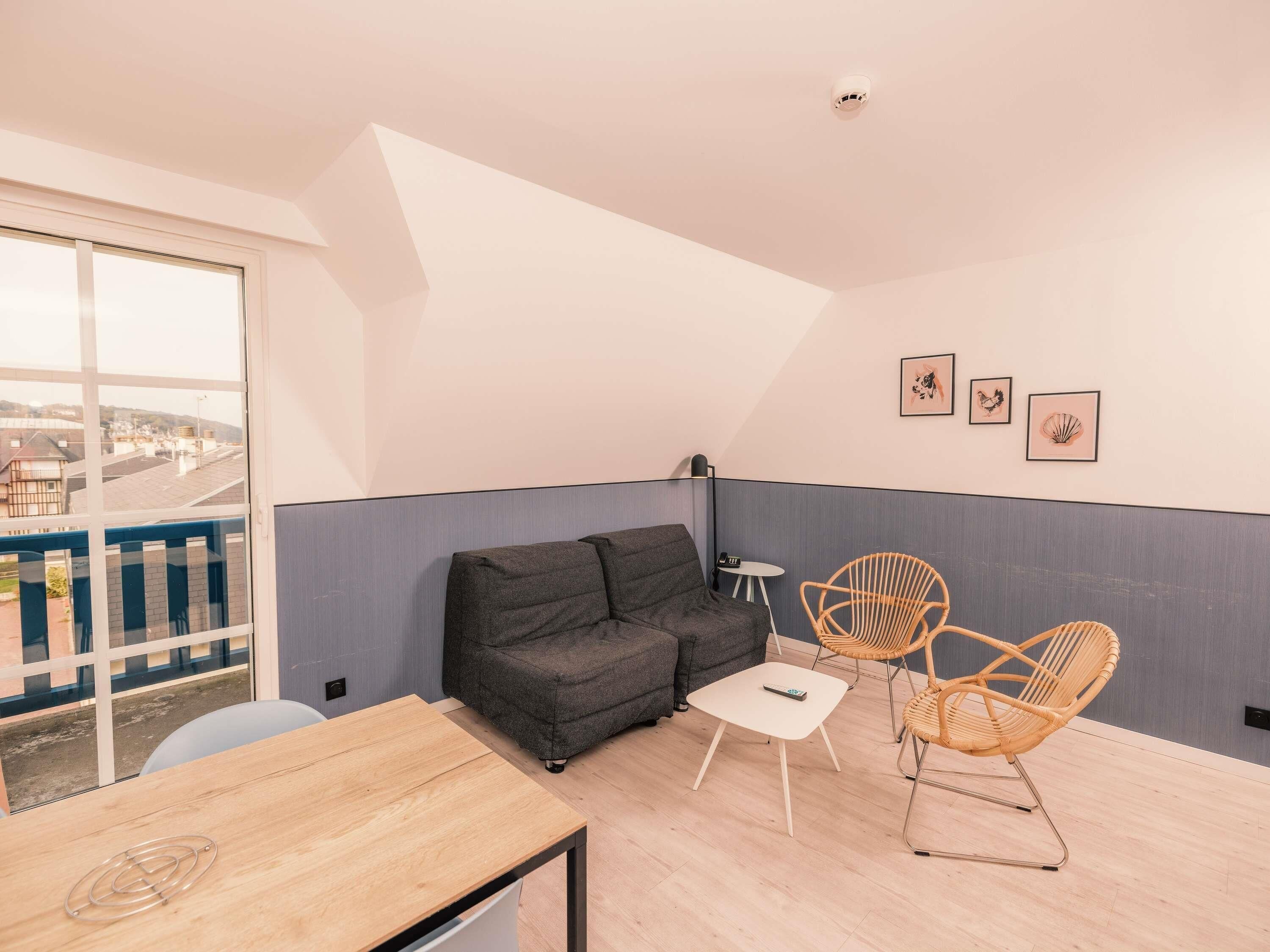 Un salon moderne avec un canapé, deux fauteuils en osier, une petite table et une vue dégagée sur l’extérieur grâce à une grande fenêtre.