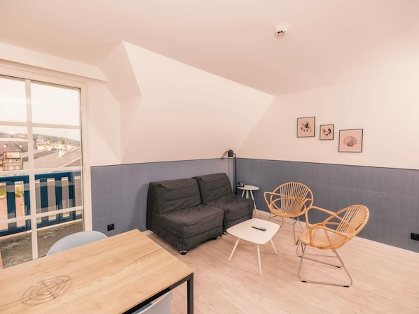 Un salon moderne avec un canapé, deux fauteuils en osier, une petite table et une vue dégagée sur l’extérieur grâce à une grande fenêtre.