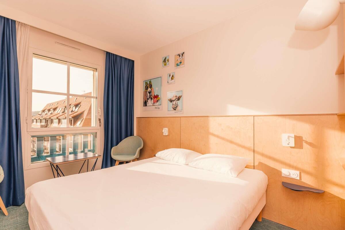 A hotel room with a bed, a chair, a window with curtains, and wall art.