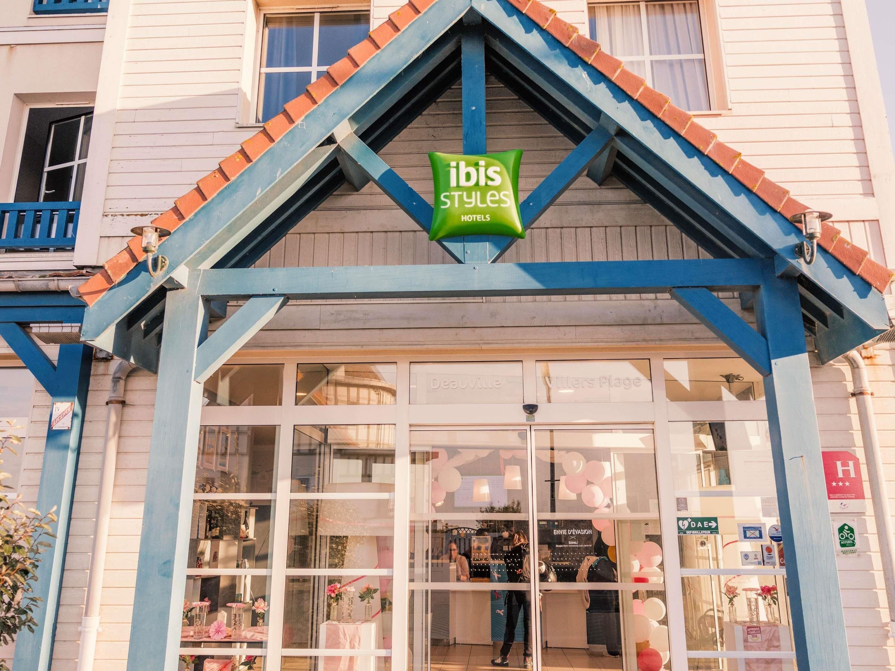 A hotel entrance with a green sign reading 'ibis Styles Hotels,' a glass facade, and a blue and red roof.