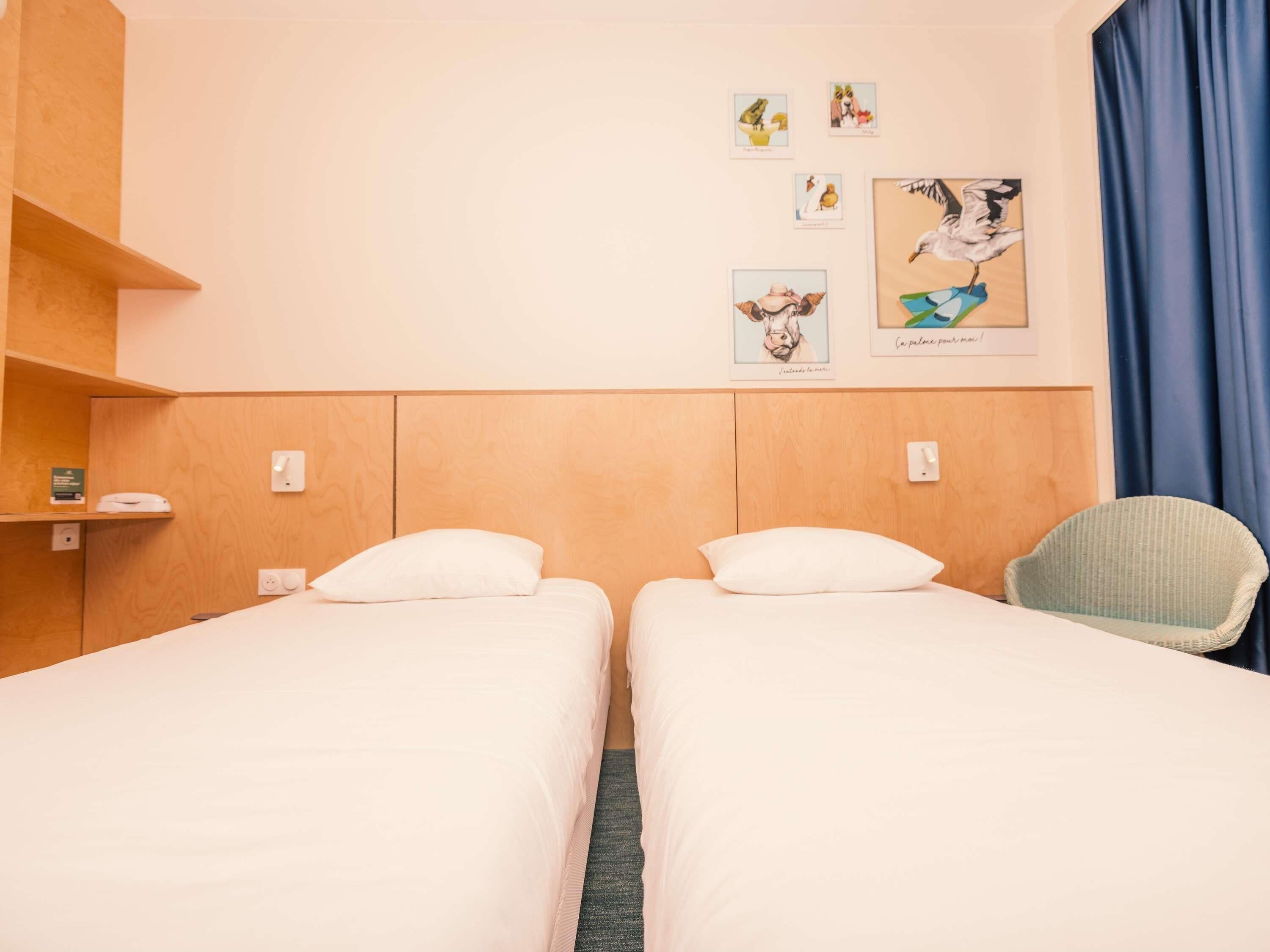 Habitación de hotel con dos camas individuales, una silla, un estante con un libro y una obra de arte en la pared.