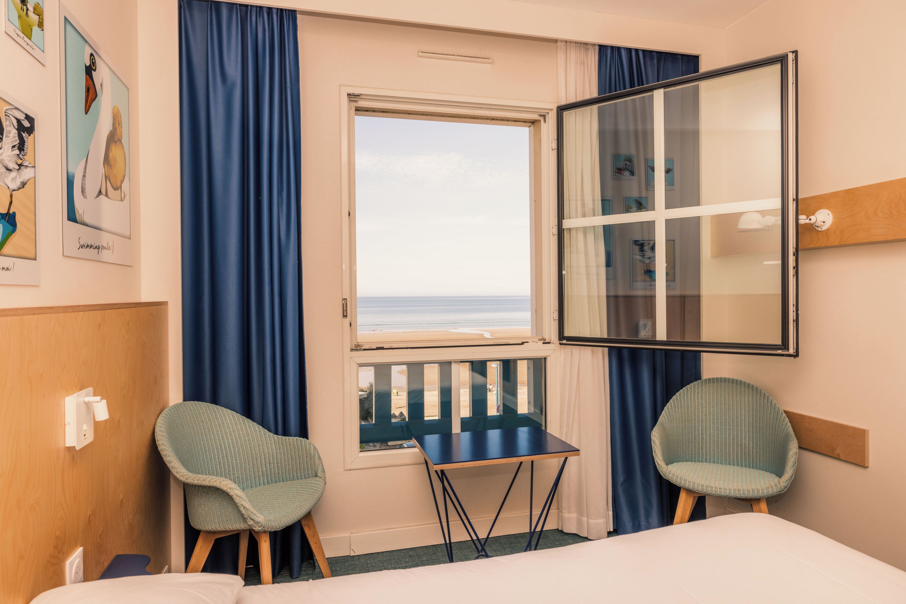 Habitación de hotel con cama, dos sillas, una mesita, ventana con vista a la playa y una obra de arte en la pared.