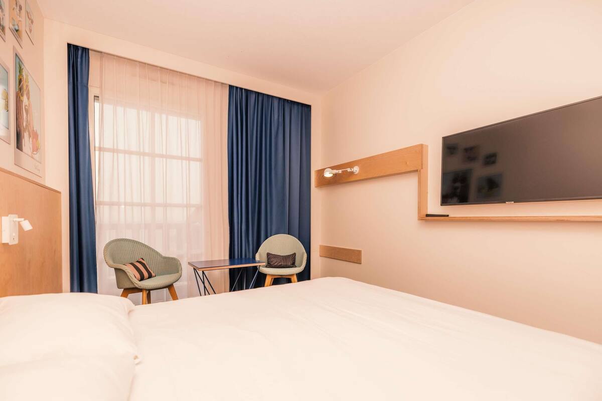 A hotel room with a bed, two chairs, a desk, a TV mounted on the wall, and a window with curtains.